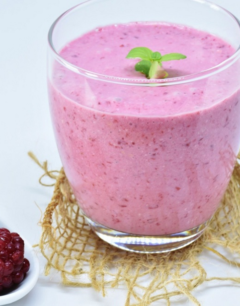 Rosa Beeren-Smoothie im Glas, dekoriert mit Minzblatt, daneben Löffel mit Brombeeren auf Jutetuch.