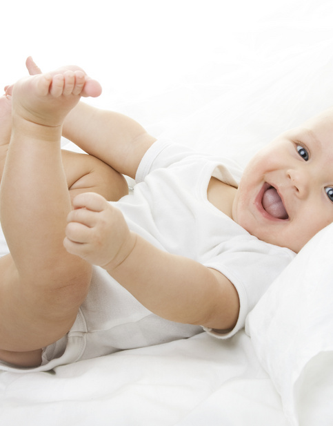 Lächelndes Baby in weißem Strampler spielt auf weißer Bettwäsche.