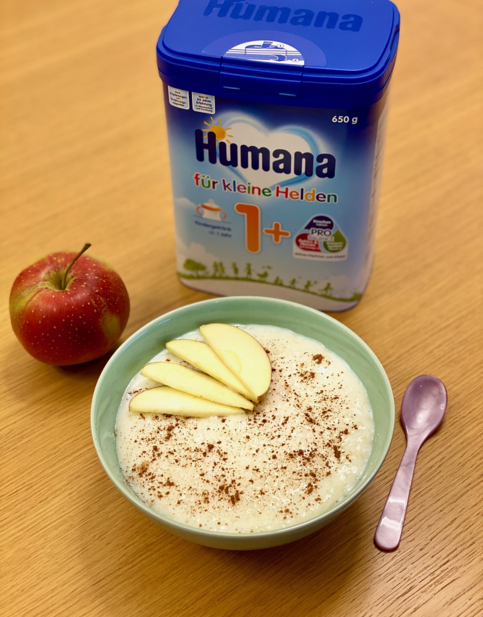 Schale mit Milchreis, bestreut mit Zimt und dekoriert mit Apfelscheiben, neben einer Packung Humana Kindergetränk und einem roten Apfel auf einem Holztisch.