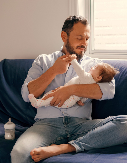 Vater sitzt auf einem Sofa und hält sein Baby im Arm, das ihn lächelnd ansieht und seinen Finger hält, neben einer Babyflasche.